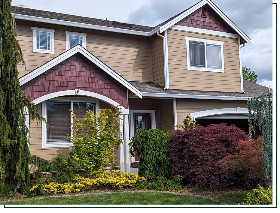A two-story suburban house with tan horizontal siding, dark red shingle accents, white trim, and a front garden